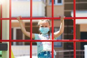 Little girl wearing a medical mask behind bars on a playground photo