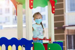 Little girl 7 years old plays in a medical mask on the playground photo