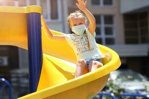 un pequeño niña en un médico máscara es montando un diapositiva en el patio de recreo foto