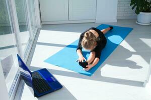 Fit little girl doing yoga and watching online lessons on laptop, exercising in the room. photo