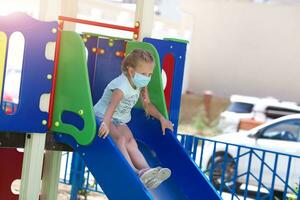 A little girl in a medical mask is riding a slide  on the playground photo