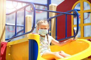 A little girl in a medical mask is riding a slide  on the playground photo