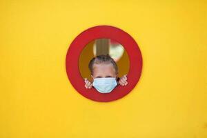 A little girl in a medical mask on her face looks out the window at the yellow playground. photo