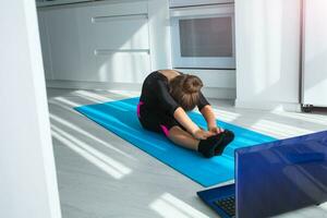 ajuste pequeño niña haciendo yoga y acecho en línea lecciones en computadora portátil, hacer ejercicio en el habitación. foto