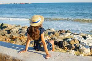 hermosa niña en un sombrero se sienta cerca el mar en piedras foto