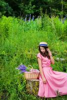 hermosa niña en un hermosa vestir se sienta en un tocón en el bosque, un ramo de flores de altramuces en un cesta foto