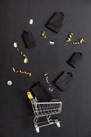 Shopping bags and shopping cart with festive decorations on black background flat lay, top view. Black friday sale concept. photo