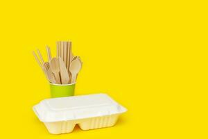 disposable wooden spoons, forks and knives in a paper cup with a corn raw lunchbox container on a yellow background with copy space, recycled photo