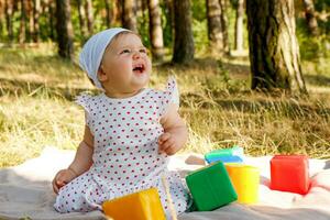 gracioso pequeño niña en un bufanda y vestir en el verano en el parque en un picnic con cubitos foto