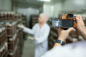 A photographer holding a digital camera on a production line is recording the work of a mushroom house worker. To be used for public relations and further marketing photo