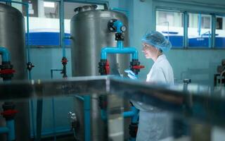 Young woman researcher in white coat and blue hat working at drinking water factory photo