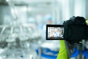 A photographer holding a digital camera on a production line is recording the work of a production line worker. To be used for public relations and further marketing photo