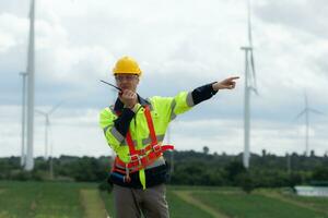 Engineer in a yellow helmet and reflective vest on the background of wind turbines using walkie talkie for communication photo