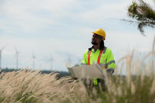 Front view of an engineer wearing a safety vest and a yellow helmet while standing in a field with wind turbines in the background photo