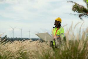 Front view of an engineer wearing a safety vest and a yellow helmet while standing in a field with wind turbines in the background photo