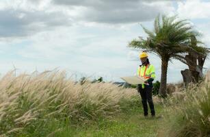 Front view of an engineer wearing a safety vest and a yellow helmet while standing in a field with wind turbines in the background photo