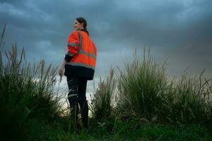 joven mujer en ingeniero uniforme y alto visibilidad con elevado brazos en pie en herboso campo a atardecer, el concepto de relajarse hora después trabajo foto