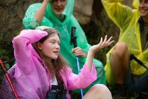 Group of friends in raincoat with backpacks hiking in the forest, Sit and shelter from the rain photo