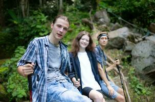 grupo de amigos con mochilas y palos sentado en un caído árbol, tomar un descanso durante el caminata foto