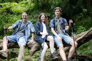 grupo de amigos con mochilas y palos sentado en un caído árbol, tomar un descanso durante el caminata foto