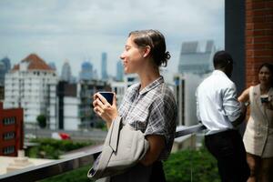 retrato de un joven mujer de negocios participación un taza de café mientras en pie en el balcón con su colegas en el antecedentes foto