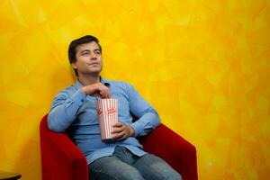 joven hombre comiendo palomitas de maiz mientras sentado en un rojo Sillón en contra un amarillo antecedentes esperando para acecho un película. foto