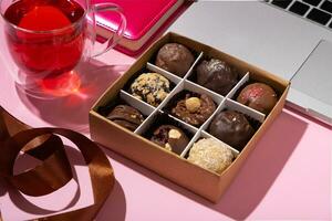 Gift box with craft chocolate candies and cup of tea on pink work desktop with laptop photo