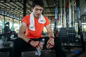 Asian bodybuilder man is sitting on bench and drinking water to relax after workout in gym. photo