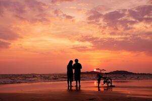 silueta Pareja en pie en el playa con un bicicleta a puesta de sol foto