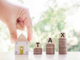 Hand holding miniature house, Wooden blocks with the word TAX on stack of coins. The concept of paying tax for house or property photo