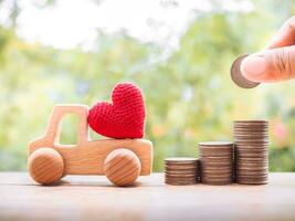 Close up of hand putting coins in stack of coins, red heart on wooden toy car for transport and manage success business concept. photo