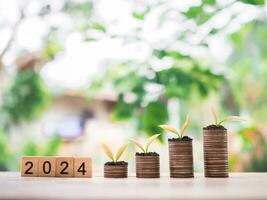 Plants growing up on stack of coins and wooden blocks with the number 2024. The concept of save money for prepare in new year 2024. photo