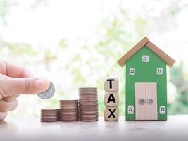 Close up hand putting coins in stack of coins, Wooden blocks with the word TAX and miniature house. The concept of Property investment, House mortgage, Real estste. photo