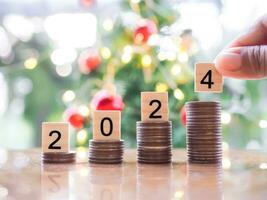 Close up hand putting number 2024 on stack of coins with Christmas light bokeh background. Merry Christmas and Happy New Year, The concept of save money for prepare in new year 2024. photo