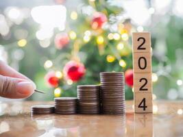 Close up hand putting coin to stack of coins and number 2024 with Christmas light bokeh background. Merry Christmas and Happy New Year, The concept of save money for prepare in new year 2024. photo