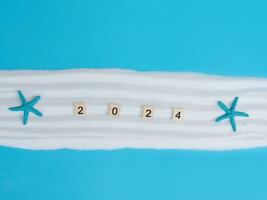 Wooden block with number 2024, seashells, starfish and white sand on blue background. Top view with copy space of summer background. The concept for summer of new year 2024 photo