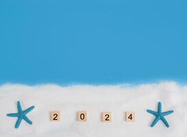 Wooden block with number 2024, seashells, starfish and white sand on blue background. Top view with copy space of summer background. The concept for summer of new year 2024 photo