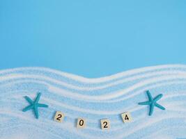 Wooden block with number 2024, seashells, starfish and white sand on blue background. Top view with copy space of summer background. The concept for summer of new year 2024 photo