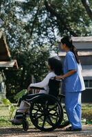 Asian young woman nurse at nursing home take care disabled senior man. Caregiver doctor serve physical therapy for older elderly patient to exercise and practice walking on walker or cane at backyard. photo