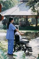 Asian young woman nurse at nursing home take care disabled senior man. Caregiver doctor serve physical therapy for older elderly patient to exercise and practice walking on walker or cane at backyard. photo