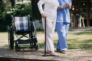 Asian young woman nurse at nursing home take care disabled senior man. Caregiver doctor serve physical therapy for older elderly patient to exercise and practice walking on walker or cane at backyard. photo