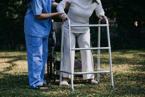 Asian young woman nurse at nursing home take care disabled senior man. Caregiver doctor serve physical therapy for older elderly patient to exercise and practice walking on walker or cane at backyard. photo