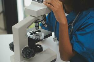 Medical worker in lab coat and sterile mask, doing a microscope analysis while her colleague are working behind photo