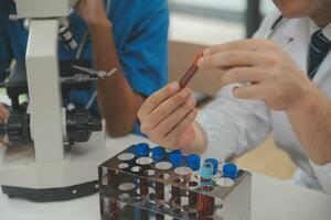 Medical worker in lab coat and sterile mask, doing a microscope analysis while her colleague are working behind photo