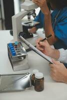 Medical worker in lab coat and sterile mask, doing a microscope analysis while her colleague are working behind photo