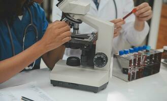 Medical worker in lab coat and sterile mask, doing a microscope analysis while her colleague are working behind photo