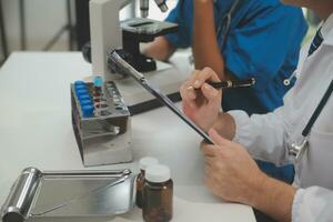 Medical worker in lab coat and sterile mask, doing a microscope analysis while her colleague are working behind photo