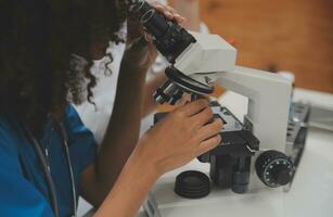 Medical worker in lab coat and sterile mask, doing a microscope analysis while her colleague are working behind photo