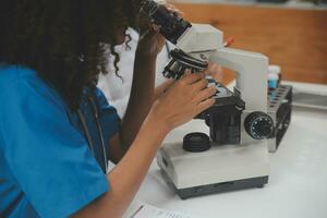 Medical worker in lab coat and sterile mask, doing a microscope analysis while her colleague are working behind photo