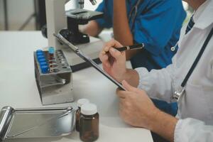 Medical worker in lab coat and sterile mask, doing a microscope analysis while her colleague are working behind photo